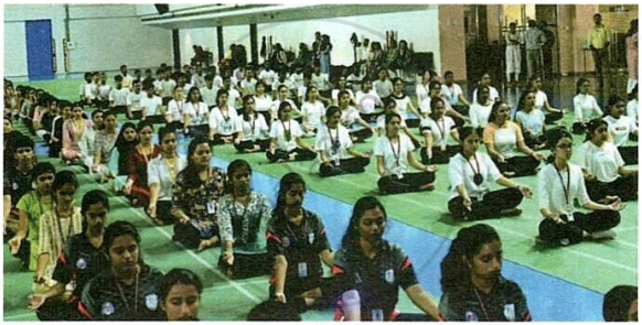 The image displays a photograph of a large group of people seated on the floor in a spacious indoor hall, participating in a mass yoga or meditation session.