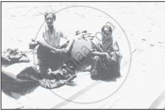 The image depicts a man and a woman sitting on the ground outdoors, engaged in selling or displaying their wares on a makeshift sheet.