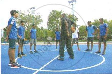 The image displays an educational photograph of a coach standing in the center circle of a sports court while conducting a training session with a group of young athletes.