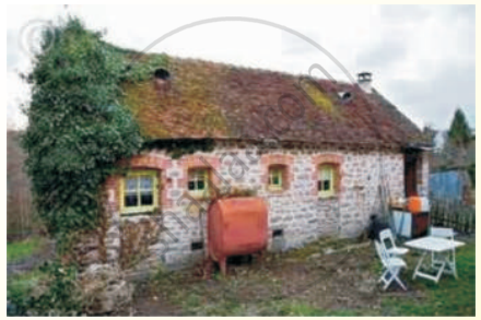 The image shows a quaint old stone cottage with a tiled roof, surrounded by greenery and simple outdoor furniture.